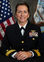 Official Photo for RDML Lacore, sitting in black Navy uniform with hands crossed in front. Behind her is the U.S. flag and another unknow official flag.
